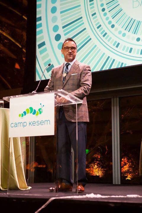 Ron Glickman speaking at a Camp Kesem podium, the camp's caterpillar logomark visible on the lectern sign.
