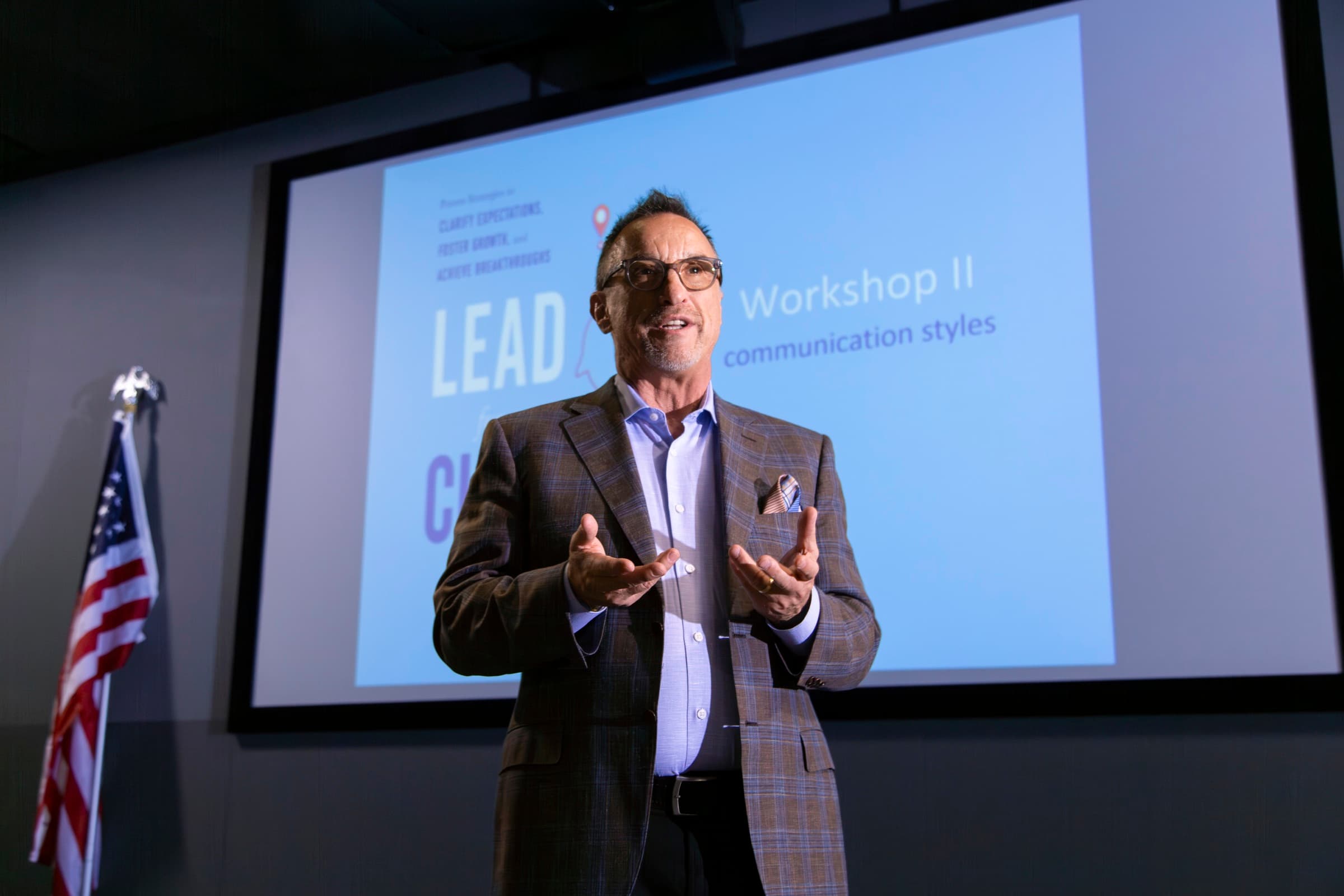 Ron Glickman on stage mid-keynote, gesturing to the room, a 'Lead for a Change' workshop slide behind him.